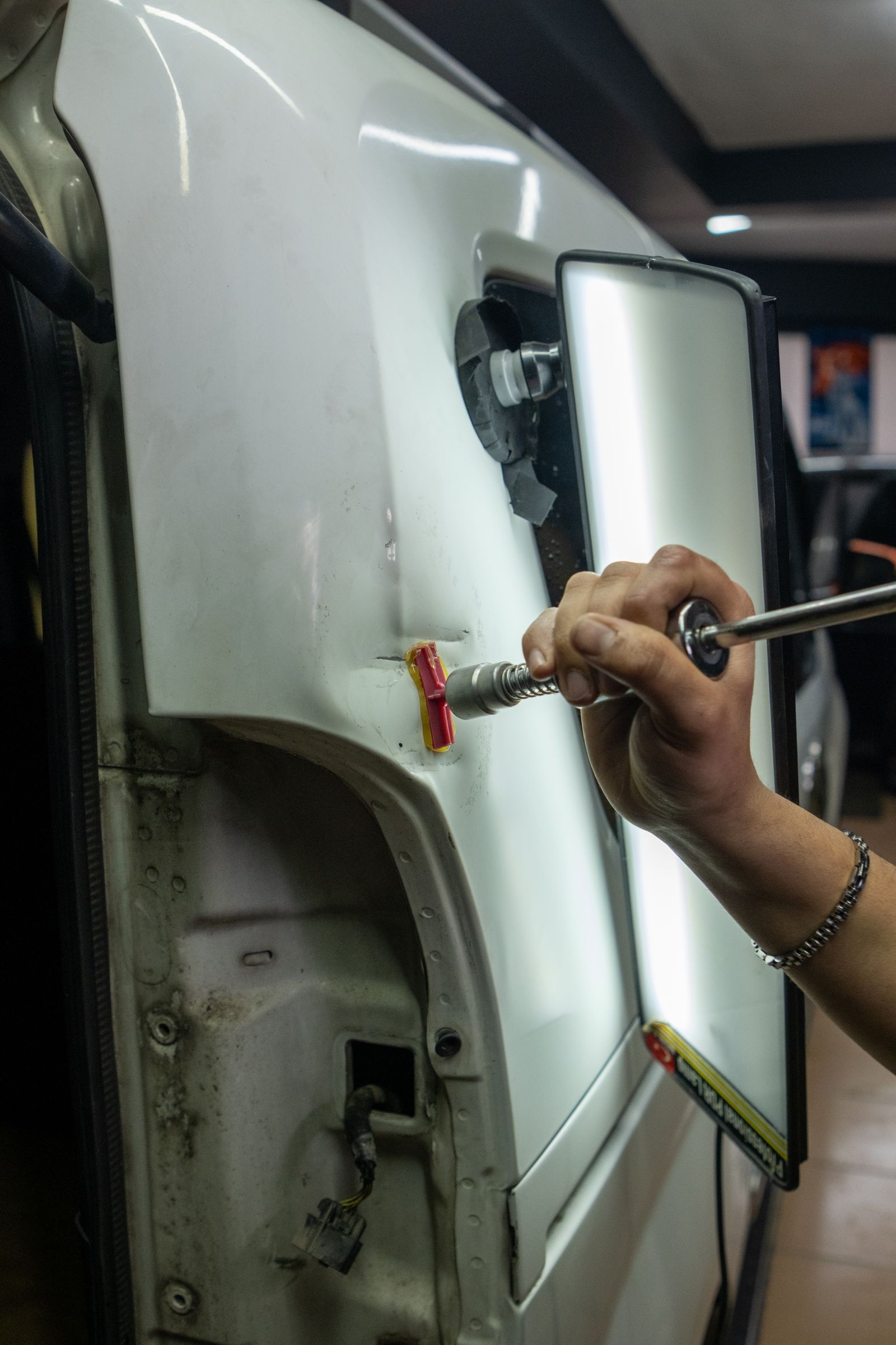 A hand using a dent-pulling tool with a red-and-yellow glue tab on the side panel of a white vehicle, with a reflective light board beside it.