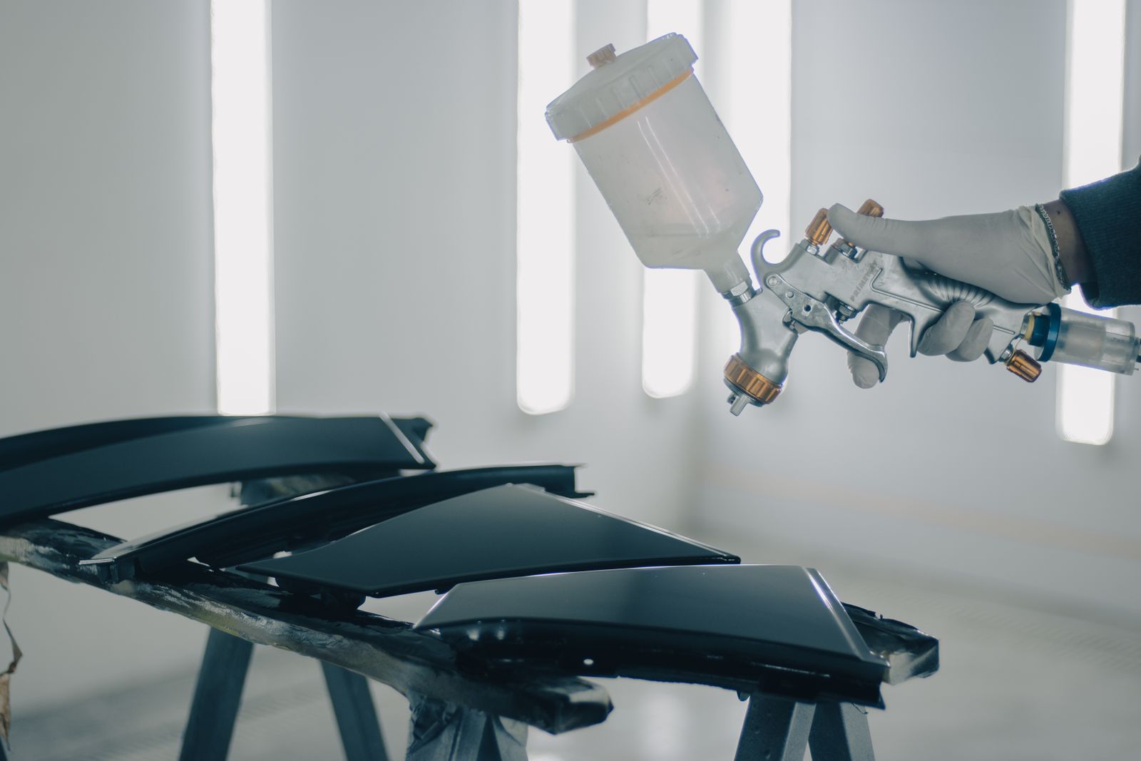 A gloved hand holding a spray gun in a bright white paint booth, beside dark glossy car body panels resting on trestles.