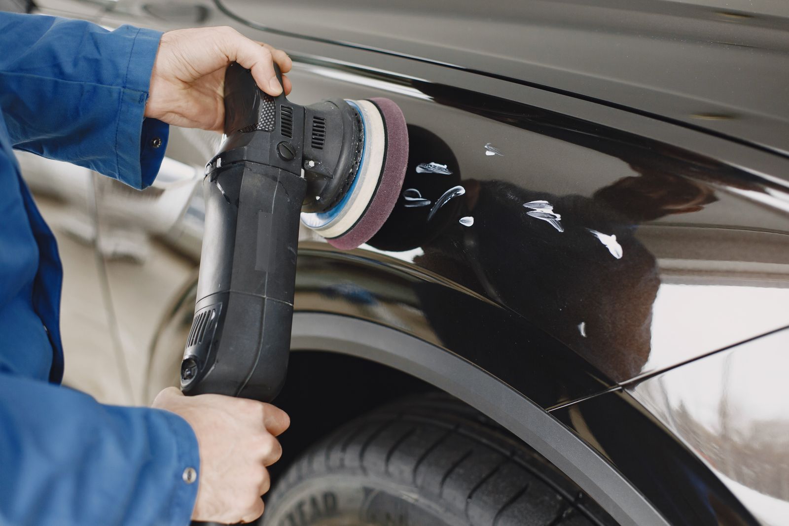 Two hands in blue overalls holding a dual-action polisher with a maroon pad on the wing of a black car, with white polish marks on the paint and part of a tyre below.